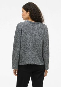 Frau mit lockigem Haar, die einen grauen Strickpullover und schwarze Hosen trägt, mit dem Rücken zu einem schlichten weißen Hintergrund.