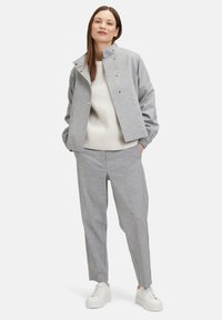 Lichtgrijze tweedelige outfit met een boxy jas met een hoge kraag en drukknoopsluiting, gecombineerd met ontspannen pasvorm broek en witte sneakers.