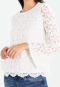 Blouse en dentelle blanche avec des manches longues évasées, un motif floral, un ourlet festonné et une texture semi-transparente, associée à un denim bleu.