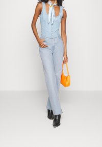 Mono de mezclilla azul claro sin mangas, con cierre de botones en la parte delantera, combinado con bolso naranja y botines negros con tachuelas.