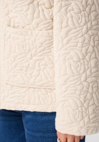 Gequilte crèmekleurige jas met een textuurpatroon, een voork pocket en een losse pasvorm met een brede manchette. Gepaard met blauwe jeans.