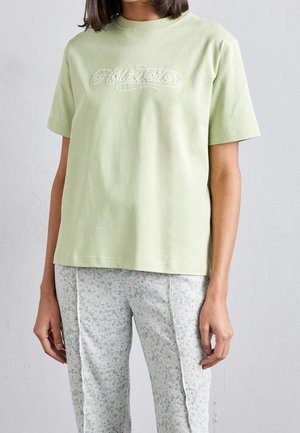 T-shirt en coton vert clair à manches courtes et col rond, arborant un texte brodé dans une police décorative sur le devant.