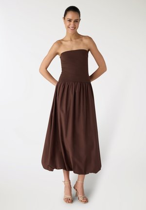 Donna sorridente con abito midi marrone senza spalline e sandali beige con tacco, in piedi con le mani dietro la schiena su sfondo bianco.