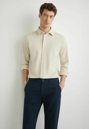 Młody mężczyzna stoi z rękami w kieszeniach, ubrany w kremową koszulę na długi rękaw z guzikami oraz ciemnogranatowe spodnie, na białym tle.