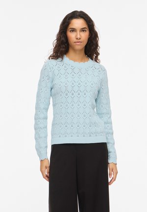 Femme aux cheveux longs et bouclés portant un pull en maille bleu clair à motifs et un pantalon noir à jambes larges, debout devant un fond blanc.