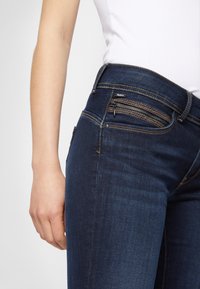 Mörkblå denimjeans med medelhög midja, dragkedjeförsedd framficka, kontrasterande sömmar och åtsittande silhuett. Slät textur.