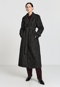 Trench coat lungo nero con trama grigia, dotato di cintura, tasche laterali, ampio colletto e taglio al ginocchio, indossato sopra pantaloni marroni e scarpe.