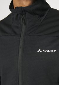 Giacca softshell nera con alto colletto, zip frontale e logo sul petto. Texture liscia con dettagli delle cuciture diagonali.