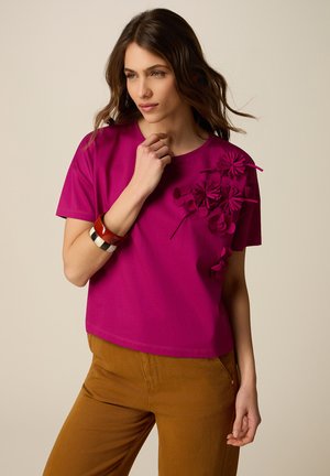 Donna che indossa una maglietta a maniche corte magenta con dettagli floreali 3D sulla spalla e pantaloni marroni, posando con una mano vicino al viso.