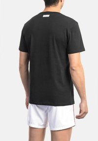 Zwarte katoenen t-shirt met een ronde hals, korte mouwen en een grijs label aan de achterkant. Het model draagt witte shorts met rode accenten.