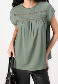 Frau trägt eine salbeigrüne Bluse mit kurzen Ärmeln, dekoriert mit Spitzenpaneelen und gewellten Kanten, hält eine schwarze Ledertasche und kombiniert sie mit schwarzen Hosen.