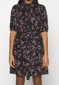 Černé květinové šaty s krátkými nadýchanými rukávy, zapínáním na knoflíky a odpovídajícím páskem ze stejné látky. Celoplošné barevné květinové vzory.