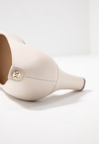 Talons hauts en cuir beige avec un bout arrondi et un design épuré. Présente un bouton accent en or et des détails de couture visibles sur le talon.