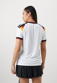 Femme portant un maillot de sport blanc avec des bandes noires, rouges et jaunes sur les épaules et une jupe plissée noire, vue de dos.