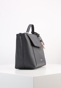 Grå læderhåndtaske med topstrop, klaplukning, lodret lynlåsdækning og en dekorativ lyserød detalje. Glat tekstur, struktureret form.