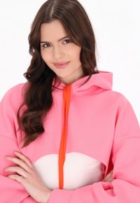 Sudadera con capucha rosa que presenta una cremallera naranja, un panel frontal blanco y una textura suave. Diseñada con un ajuste relajado y mangas amplias.