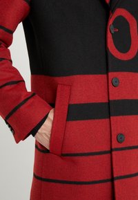 Czerwono-czarny płaszcz z mieszanki wełny z poziomymi paskami, wyciętym kołnierzem, boczną kieszenią oraz czarnymi guzikami z czerwonym przeszyciem.