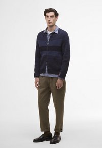 Marineblauwe gebreide cardigan met textuurstrepen, een voorrits en een lusdetail. Draagt over een lichtblauw shirt en gecombineerd met olijfgroene broeken en zwarte schoenen.