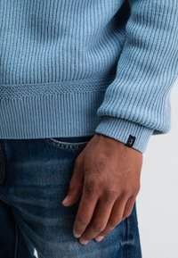 Hellblauer, gerippter Strickpullover mit strukturiertem Bündchen und schwarzem Etikett. Getragen zu dunkelblauen Jeans, wobei eine Nahaufnahme des Ärmels und der Hand gezeigt wird.