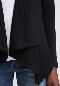 Musta, kevyt kietaisumallinen neuletakki pehmeästä kankaasta; siinä on epäsymmetrinen helma ja pitkät hihat, kerrostettuna vaalean topin päälle.