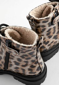 Bottes cheville en imprimé léopard avec doublure en peluche beige, fermeture éclair noire et semelles épaisses en caoutchouc noir sur un fond blanc.