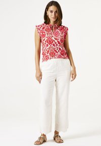 Blusa sin mangas roja con patrón floral y lazo en el cuello, combinada con pantalones anchos blancos bordados y sandalias planas.