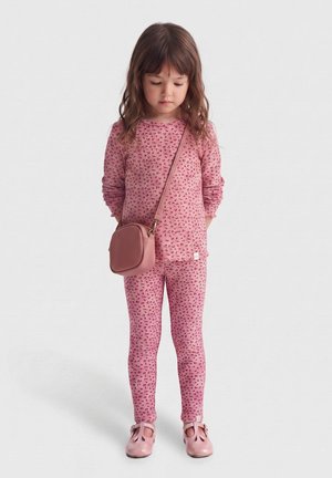 Jeune fille vêtue d’un haut à manches longues rose à fleurs et de leggings, avec des chaussures roses et un petit sac bandoulière, debout la tête baissée.
