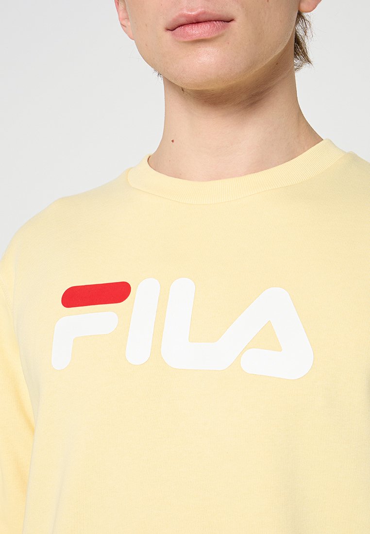 Sudadera de color amarillo claro con un gran logo blanco de FILA, acentuado con una línea roja. La tela es suave y lisa con un cuello redondo estándar.