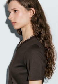 Jonge vrouw met lang golvend bruin haar draagt een zwart T-shirt met korte mouwen en een dikke zilveren ketting, kijkend naar links tegen een lichte achtergrond.