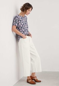 Blusenshirt mit floralen Druck und kurzen Ärmel, kombiniert mit weißen, weiten Hosen und braunen, geflochtenen Lederschuhen. Lässiges Outfit mit Gürtel vor einer neutralen Wand.