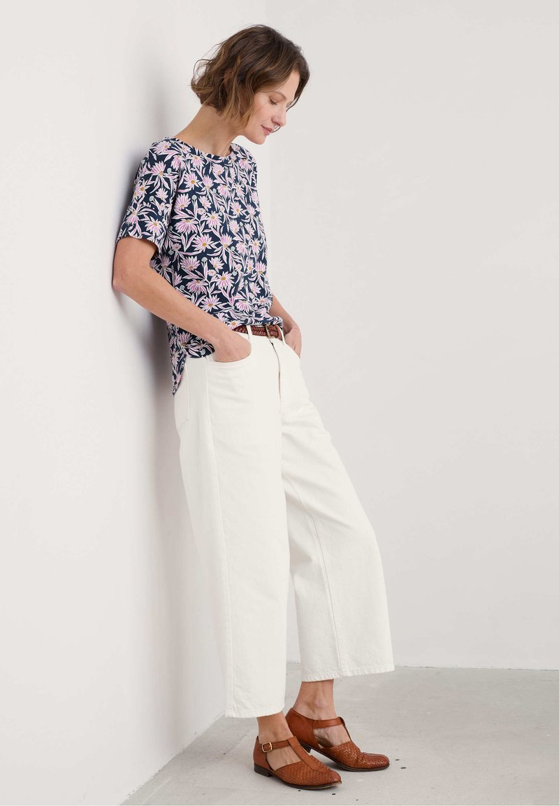 Blusenshirt mit floralen Druck und kurzen Ärmel, kombiniert mit weißen, weiten Hosen und braunen, geflochtenen Lederschuhen. Lässiges Outfit mit Gürtel vor einer neutralen Wand.