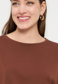 Νεαρή γυναίκα που χαμογελά, φοράει καφέ μπλούζα με στρογγυλή λαιμόκοψη και χρυσούς κρίκους στα αυτιά, με φόντο λευκό και απλό.
