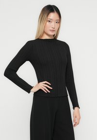 O femeie tânără cu păr drept în degrade purtând un top negru cu dungi lungi la mâneci și pantaloni negri, pozând cu o mână pe șold.