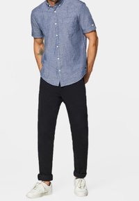 Man in een kortebloemig blauw overhemd, zwarte opgerolde broek en witte sneakers met zijn handen in zijn zakken.