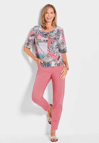 Mehrfarbige Bluse mit Schalkragen, kurzen Ärmeln und verschiedenen Mustern. Kombiniert mit rosa schmal geschnittenen Hosen und beigen Sandalen.