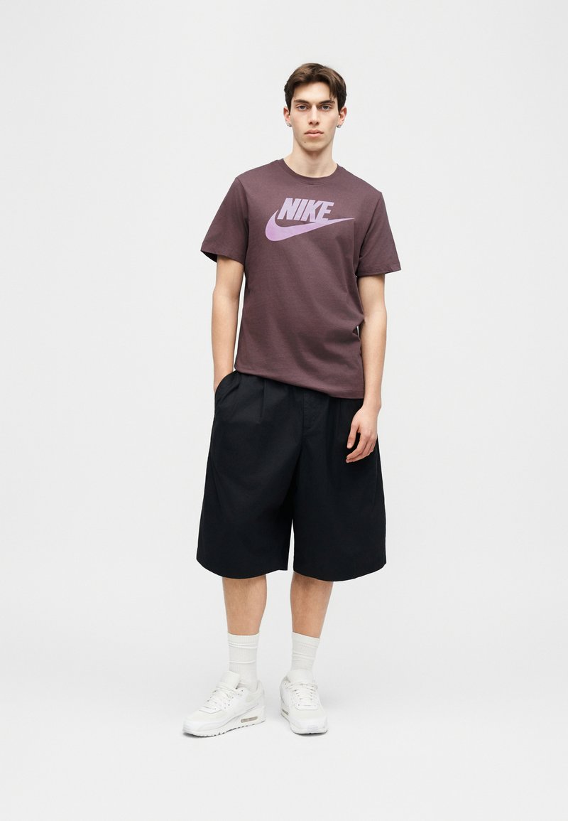 Jonge man met handen in zijn broekzakken, gekleed in een paars Nike T-shirt, zwarte wijde korte broek, witte sokken en witte sneakers, tegen een effen achtergrond.