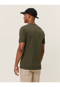 oliwkowa bawełniana koszulka z krótkim rękawem i okrągłym dekoltem, wyposażona w małe czerwone haftowane logo z tyłu, zestawiona z beżowymi spodniami.