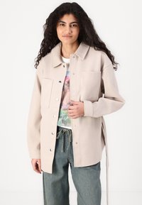 Giacca beige con bottoni in morbido tessuto, caratterizzata da una tasca sul petto, bottoni in metallo argentato e lacci laterali. Abbinata a una t-shirt grafica e jeans sbiaditi.