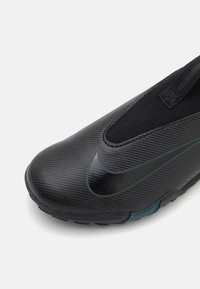 Svart atletisk sko med en strukturerad ovandel, slip-on design och gröna detaljer. Har en slät tåkappa och flexibel sula.