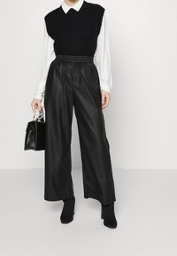 Suéter negro sin mangas de punto sobre una blusa blanca con cuello, combinado con pantalones de imitación cuero negros de pierna ancha y botas de tobillo negras con tacón.