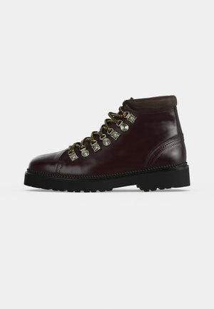 Bota de senderismo de cuero marrón oscuro con suela negra, ojales metálicos y cordones redondos amarillo-negros mostrada de lado sobre un fondo liso.