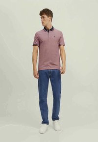 Camisa polo em borgonha com gola canelada, detalhes em azul-marinho e mangas listadas; combinada com jeans azuis e ténis brancos.