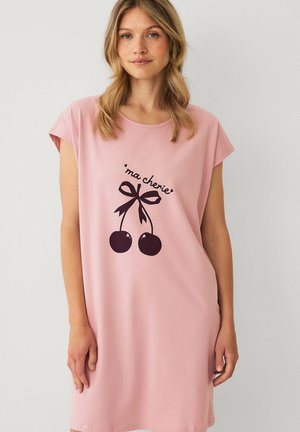 Vrouw draagt een losse roze jurk met korte mouwen en een zwart kersmotief en de tekst "ma chérie" aan de voorkant.
