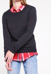 Kvinna med en texturerad svart tröja över en röd rutig skjorta, matchat med ljusblå jeans, händerna korslagda framför sig.