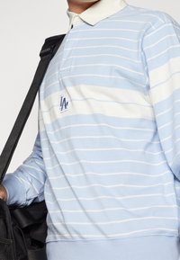 Pruhovaná polokošile s dlouhým rukávem v světle modré a bílé barvě, s límečkem a vyšitým logem na hrudi. Měkký bavlněný materiál.