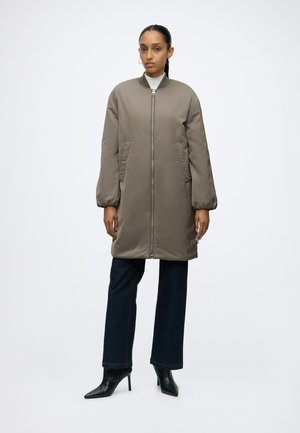 Vrouw staand en naar voren gericht, draagt een lange taupekleurige jas, donkere broek en zwarte laarzen met hak tegen een effen witte achtergrond.