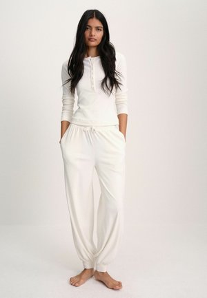 Woman standing barefoot wearing white long-sleeve henley top and matching drawstring pants against plain light background.