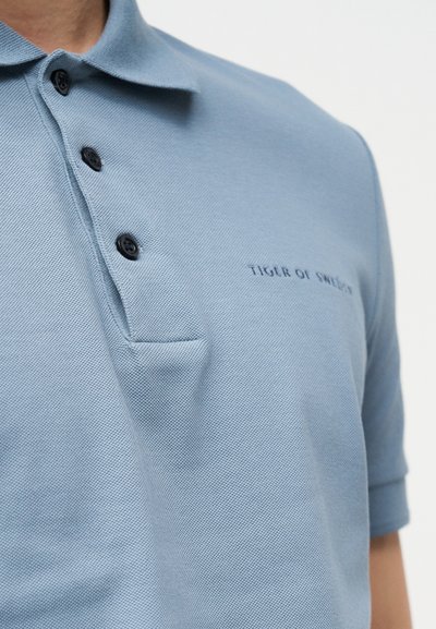 Γαλάζιο πόλο πουκάμισο με τρία μαύρα κουμπιά και κεντημένο λογότυπο "Tiger of Sweden" στο στήθος.