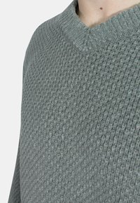 Maglione grigio lavorato a maglia con un design testurizzato e un colletto a coste. Il tessuto appare morbido con una leggera lucentezza, mostrando una vestibilità casual.