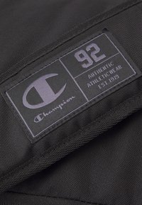 Etiqueta de tela negra con texto púrpura que dice "Champion," "92," y "AUTHENTIC ATHLETICWEAR EST. 1919," con un logotipo y detalles de costura.